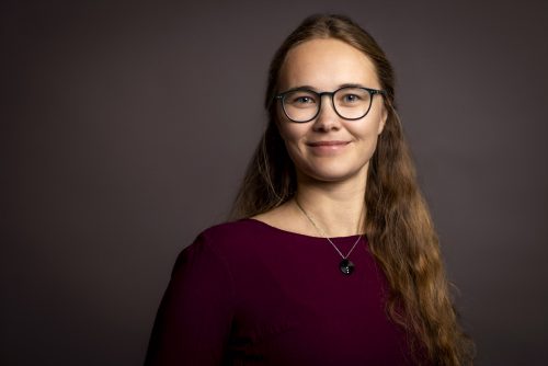 Frau mit langen, gewellten Haaren trägt ein dunkelrotes Oberteil und eine Halskette mit rundem Anhänger vor einfarbigem Hintergrund