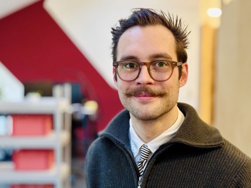 Mann mit dunklem, zurückgekämmtem Haar trägt schwarzen Reißverschluss-Pullover über weißem Hemd und gestreifter Krawatte, unscharfer Hintergrund mit Regalen und roter Wand.