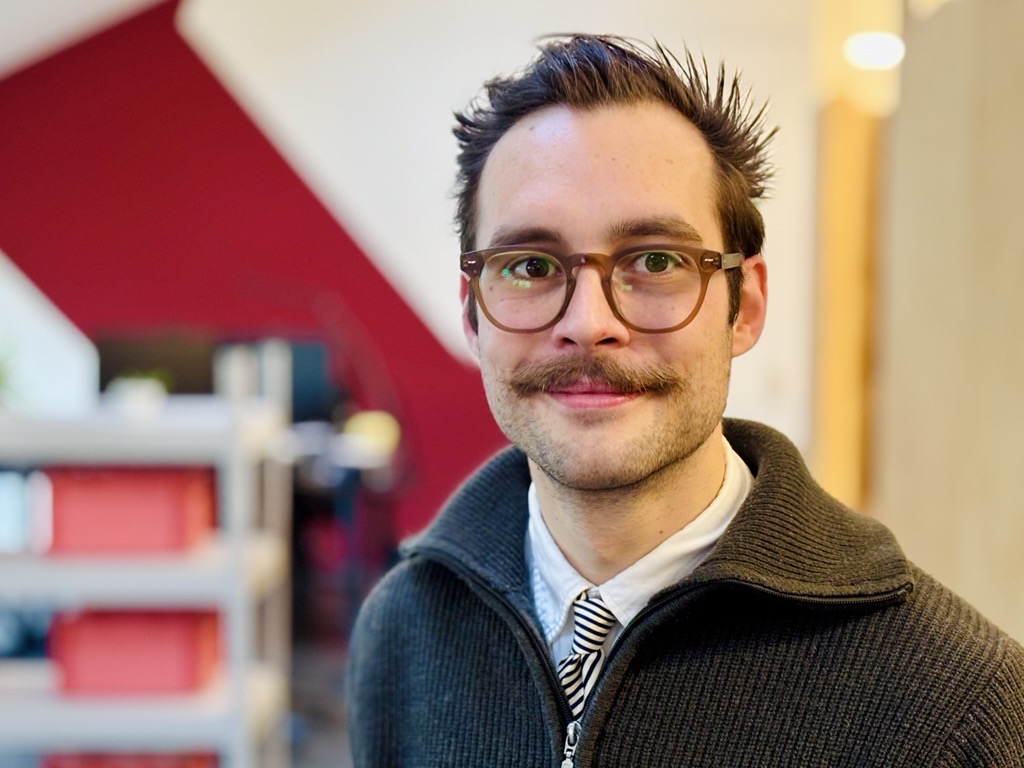 Mann mit dunklem, zurückgekämmtem Haar trägt schwarzen Reißverschluss-Pullover über weißem Hemd und gestreifter Krawatte, unscharfer Hintergrund mit Regalen und roter Wand.