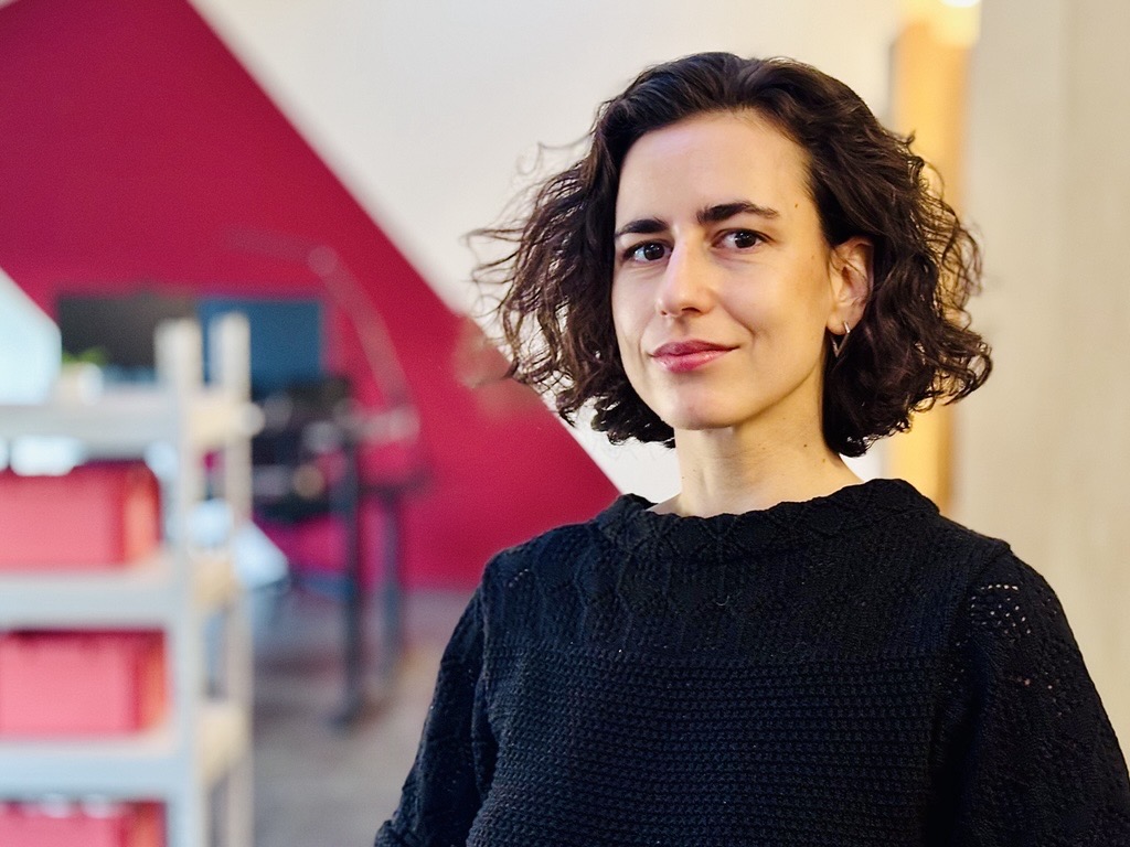 Frau mit schulterlangem, lockigem Haar trägt schwarzen Pullover und steht vor roter und gelber Wand in einem Raum mit Regalen.