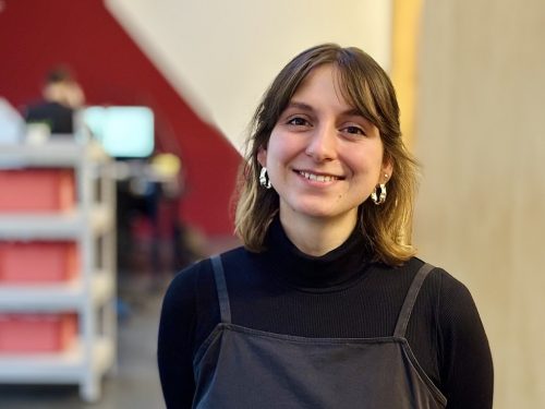 Eine junge Frau mit schulterlangem Haar lächelt in die Kamera. Sie trägt ein schwarzes Oberteil mit Trägern darüber und auffällige goldene Ohrringe. Der Hintergrund zeigt ein unscharfes Büro oder Arbeitsumfeld.