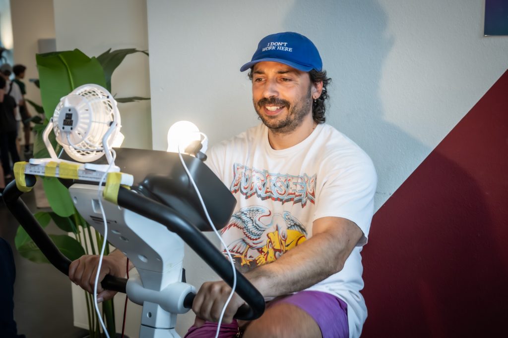 Mann mit blauer Kappe und weißem T-Shirt sitzt auf einem Heimtrainer vor zwei kleinen Ventilatoren
