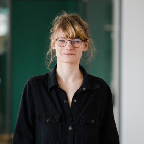 Porträtfoto einer jungen Frau mit dunkelbraunen Haaren. Sie trägt ein dunkles Hemd mit Knöpfen, lächelt freundlich in die Kamera, der Hintergrund ist grünlich.