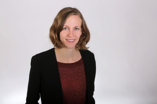 Frau mit schulterlangem, gewelltem Haar trägt schwarzen Blazer und rot gemustertes Oberteil vor weißem Hintergrund