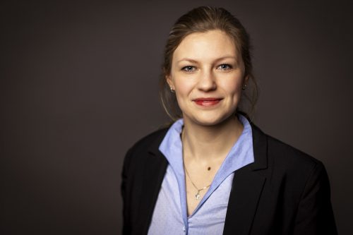 Porträtfoto einer Frau mit hellen Haaren, die zu einem Zopf zurückgebunden sind. Sie trägt ein hellblaues Hemd unter einem dunklen Blazer sowie eine Kette mit Kreuzanhänger. Sie blickt freundlich in die Kamera und lächelt leicht. Der Hintergrund ist neutral dunkel gehalten.