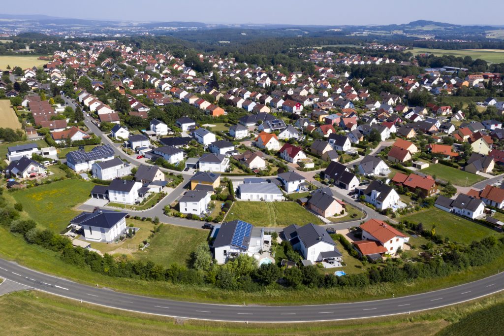 Luftaufnahme einer deutschen Vorortsiedlung mit vielen Einfamilienhäusern, Gärten und Straßen. Einige Dächer sind mit Solarpanelen ausgestattet. Im Hintergrund sind Felder, Wälder und sanfte Hügel zu sehen.