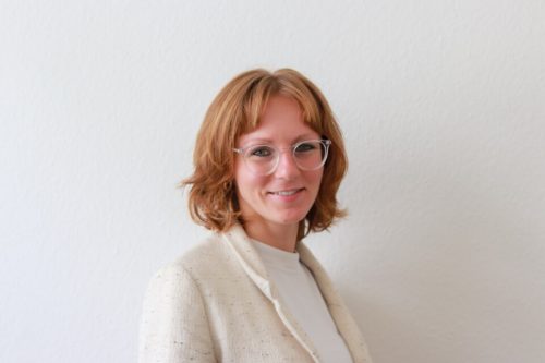 Portraitfoto einer Frau mit, weißer transparenter Brille und roten kinnlangen Haaren, vor einer weißen Wand. Sie trägt ein weißes T-Shirt und darüber ein creme Weißen Baumwollblazer.
