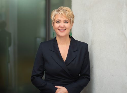 Porträt einer blondhaarigen Frau mittleren Alters mit kurzem Haar. Sie trägt einen schwarzen Hosenanzug, lehnt an einer Betonwand und lächelt in die Kamera.