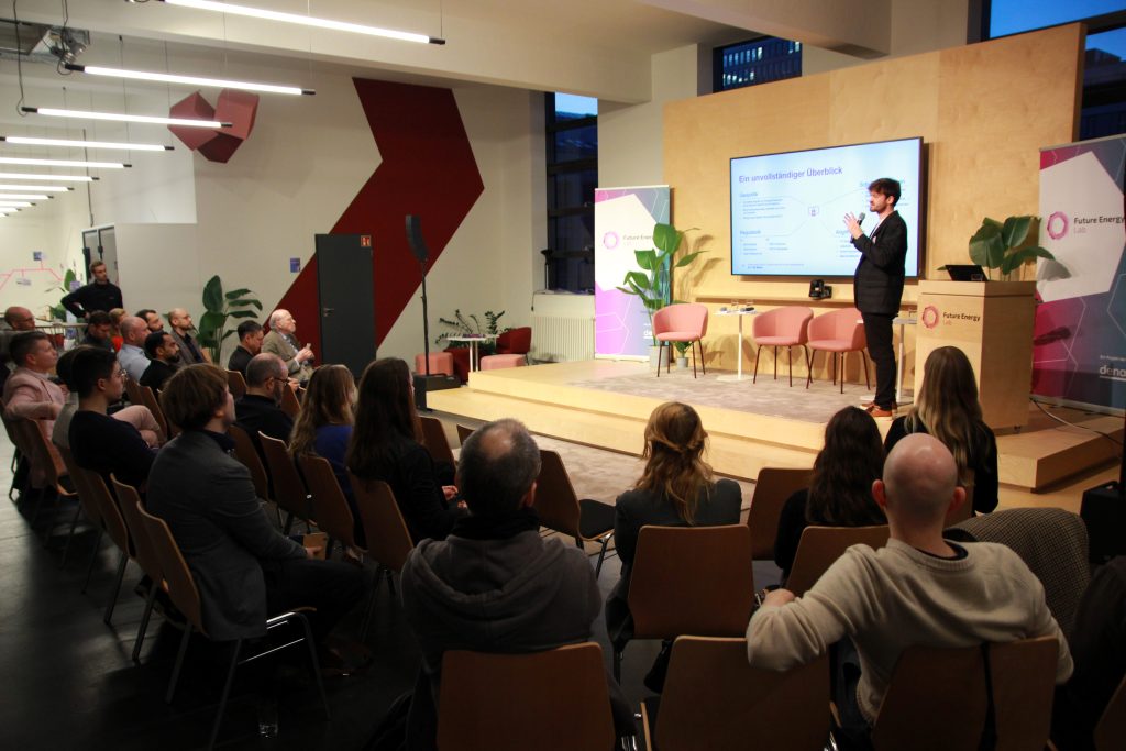 Ein Mann steht auf einer Bühne und hält einen Vortrag vor einem Publikum in einem modernen Veranstaltungsraum mit Holzverkleidung und Pflanzen.