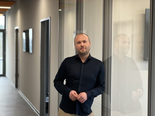 Person steht in einem modernen Büroflur neben einer Glaswand, in der sich die Person spiegelt. Die Person trägt ein dunkles Hemd und blickt leicht lächelnd in die Kamera. Der Flur ist hell und minimalistisch gestaltet.