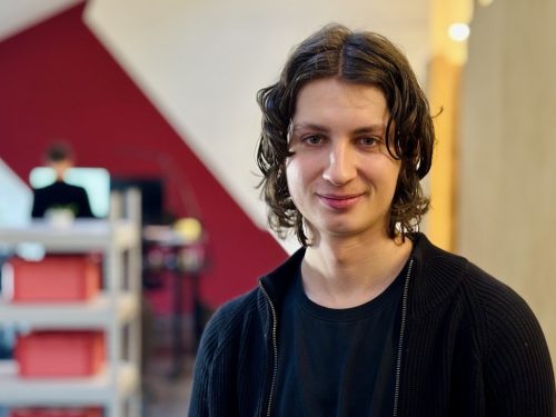 Junge Person mit schulterlangem, gewelltem Haar trägt schwarzen Pullover und T-Shirt, steht vor roter Wand und Regal mit roten Boxen.