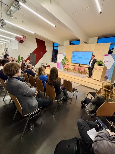 Mann hält Vortrag auf Bühne mit Bildschirm, Publikum sitzt auf Stühlen und hört zu, moderner Veranstaltungsraum mit Holzrückwand.