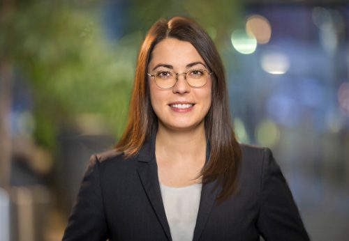 Frau mit langen braunen Haaren trägt weißen Pullover und schwarzen Blazer, unscharfer Hintergrund mit Lichtern.