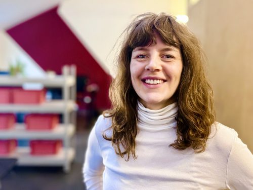 Frau mit langen, lockigen Haaren trägt weißen Rollkragenpullover und steht vor einem Regal mit bunten Boxen und einer roten Wanddekoration.