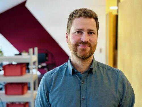 Mann mit blauem Hemd steht vor einem Regal mit roten Boxen in einem Büro mit roter und gelber Wand.
