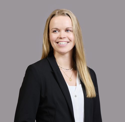 Weiblich gelesene Person mit langen blonden Haaren trägt schwarzen Blazer, weiße Bluse und zwei Ketten vor einfarbig grauem Hintergrund