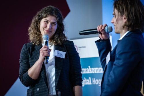 Zwei Personen im Gespräch, eine hält ein Mikrofon, die andere spricht in ein Mikrofon, im Hintergrund ein Banner mit Textfragmenten