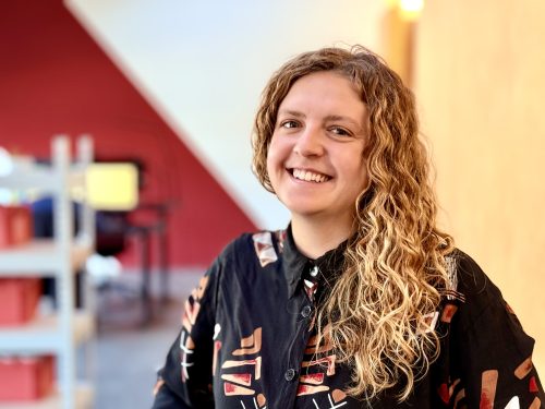 Weiblich gelesene Person mit langen, lockigen Haaren trägt ein schwarzes Hemd mit buntem Muster vor einem unscharfen Hintergrund mit roten und gelben Flächen