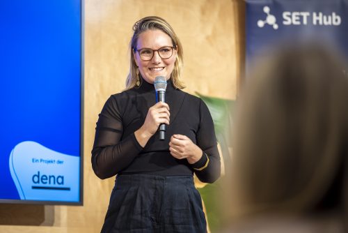 Frau mit schwarzem Oberteil und Mikrofon in der Hand steht vor einem Bildschirm mit dem Logo 'dena' und einem Schriftzug 'SET Hub'