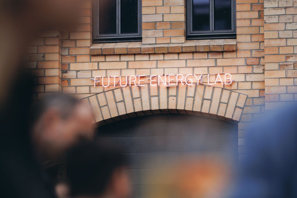 Neonlicht-Schriftzug 'FUTURE ENERGY LAB' an einer Ziegelsteinwand über einem Tor mit verschwommenen Personen im Vordergrund