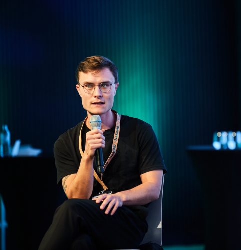 Person sitzt mit verschränkten Beinen auf einem Stuhl und hält ein Mikrofon in der rechten Hand