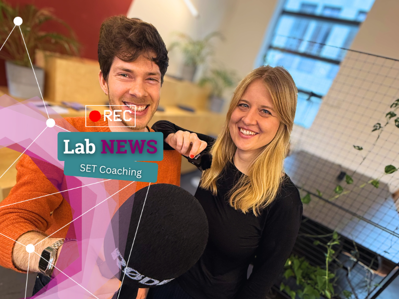 Zwei Personen sitzen nebeneinander, eine hält ein Mikrofon, im Vordergrund ein eingeblendetes rotes Aufnahme-Symbol und Textfelder mit 'Lab NEWS' und 'TAKE OVER: SET Coaching'