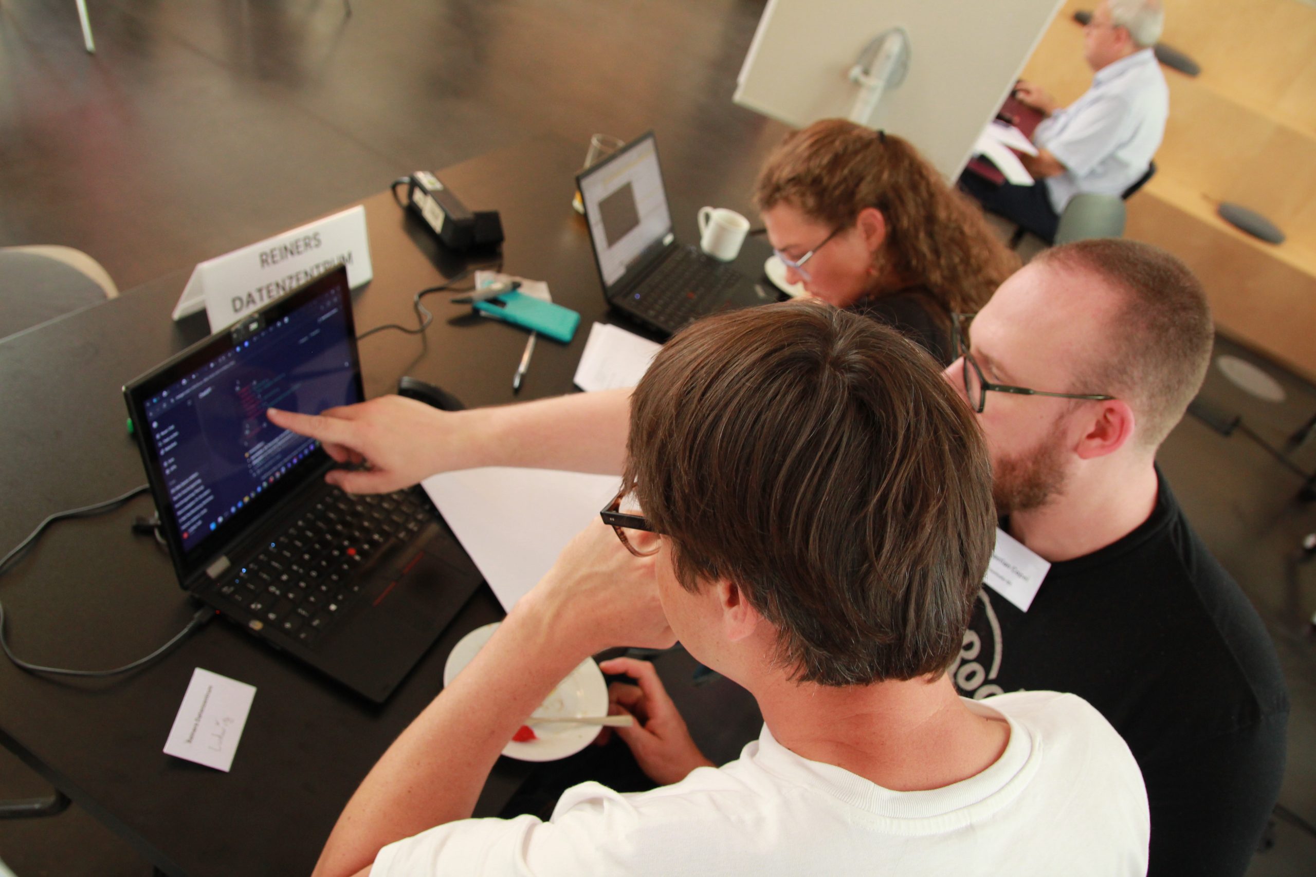 Drei Personen sitzen an einem Tisch und schauen auf einen Laptop, eine Person zeigt auf den Bildschirm
