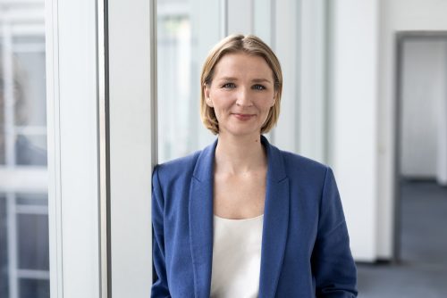 Frau mit kurzem blonden Haar trägt ein blaues Sakko und ein weißes Oberteil steht vor großen Fenstern in einem modernen Bürogebäude