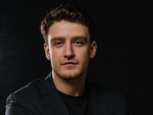 Mann mit dunklem Sakko und schwarzem T-Shirt vor schwarzem Hintergrund sitzt mit verschränkten Armen