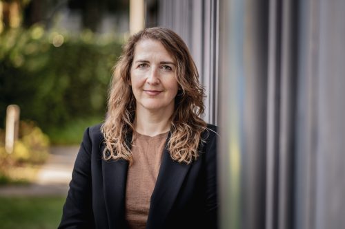 Frau mit schulterlangen gewellten Haaren trägt braunes Oberteil und schwarzen Blazer steht vor unscharfem Hintergrund und lehnt an einer Säule