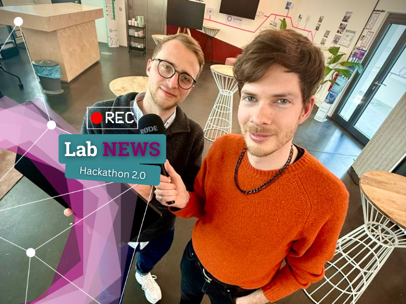 Zwei Personen in einem modernen Innenraum, eine hält ein Mikrofon mit dem Schriftzug 'Lab NEWS Hackathon 2.0', im Bild ein rotes Aufnahme-Symbol 'REC'