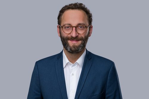 Mann mit dunklem, lockigem Haar trägt weißen Hemdkragen und dunkelblauen Anzug vor grauem Hintergrund