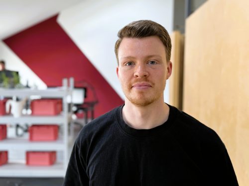 Mann mit kurzem Haar und schwarzem Pullover vor rotem und beigem Hintergrund