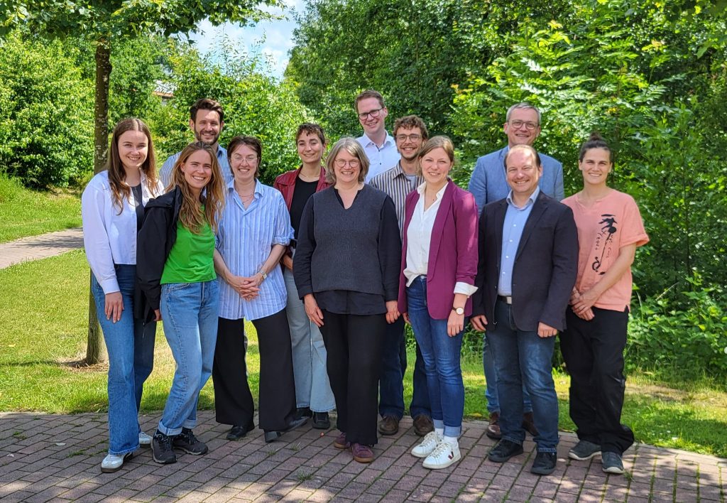 Gruppe von elf Personen steht auf gepflastertem Weg vor grünen Bäumen im Freien