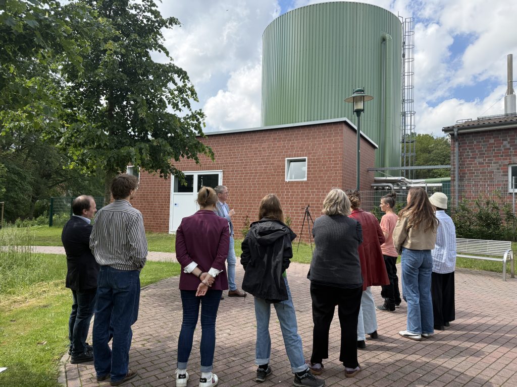 Gruppe von Menschen steht vor einem großen zylindrischen Wassertank neben einem Backsteingebäude im Freien