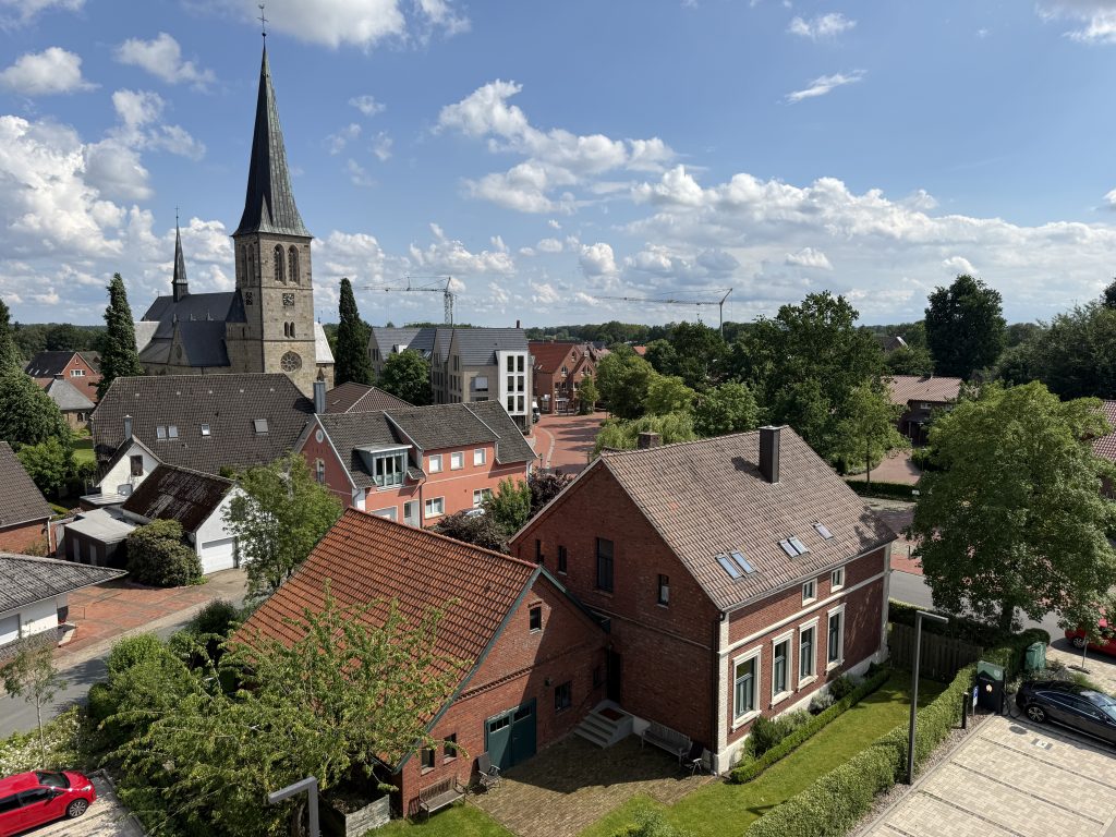 Blick über ein Dorf mit Backsteinhäusern, einer Kirche mit hohem Turm und einem Parkplatz bei sonnigem Himmel mit Wolken