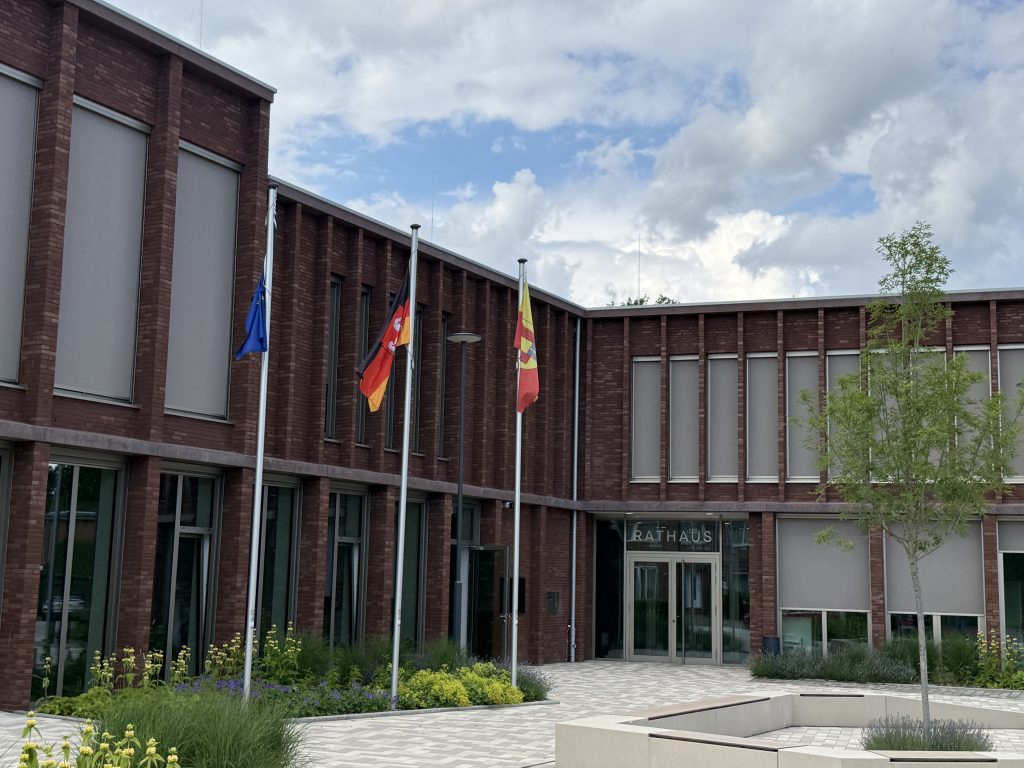 Backstein-Rathausgebäude mit drei Fahnenmasten und bepflanztem Vorplatz unter bewölktem Himmel