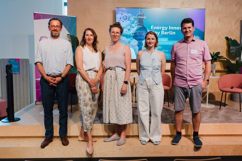 Fünf Personen stehen nebeneinander auf einer Bühne vor einem Bildschirm mit der Aufschrift 'Energy Innovation Day Berlin', sie tragen sommerliche Kleidung und lächeln in die Kamera