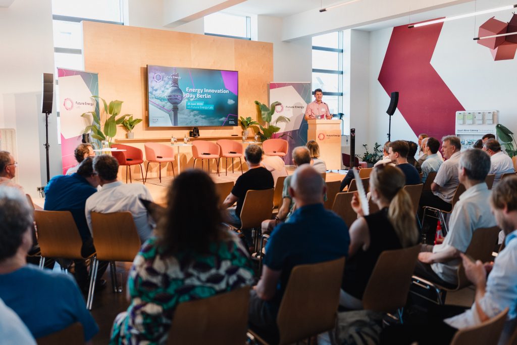 Vortragender steht an einem Rednerpult vor Publikum in einem modernen Konferenzraum mit großem Bildschirm und Pflanzen
