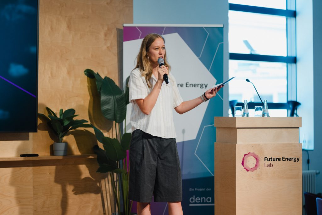 Frau steht mit Mikrofon und Tablet vor einem Rednerpult mit Aufschrift 'Future Energy Lab' und spricht in einem hellen Raum mit großen Fenstern