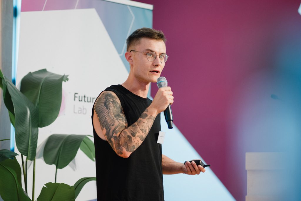 Junger Mann mit Brille und Tattoo am linken Arm hält Mikrofon und präsentiert vor einem Banner und Pflanzen