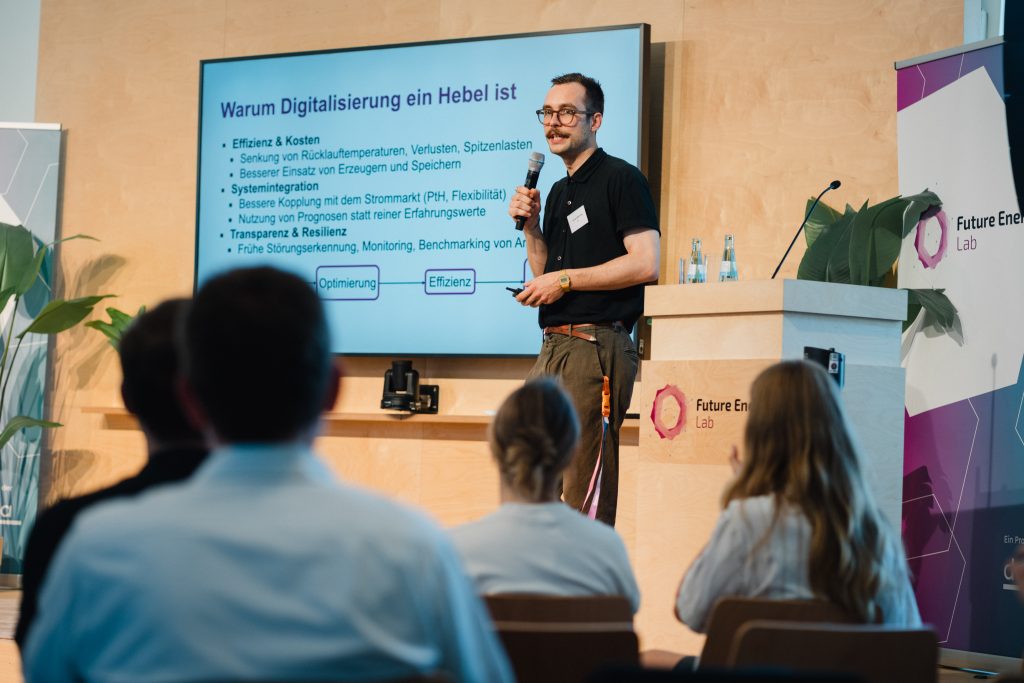 Mann mit Brille und Mikrofon hält Vortrag vor Bildschirm mit Präsentationstext und Publikum im Vordergrund