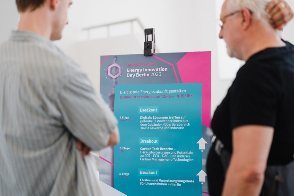 Zwei Männer betrachten ein Informationsplakat zum Energy Innovation Day Berlin 2026 mit Programmpunkten zu digitalen Energielösungen, Carbon Tech und Förderangeboten