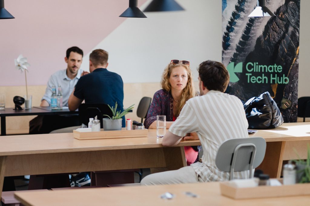 Zwei Paare sitzen an Tischen in einem hellen Raum, im Hintergrund ein Banner mit der Aufschrift 'Climate Tech Hub'