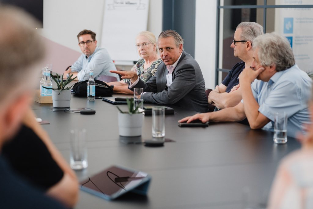 Mehrere Personen sitzen an einem langen Konferenztisch, ein Mann in der Mitte gestikuliert während einer Besprechung