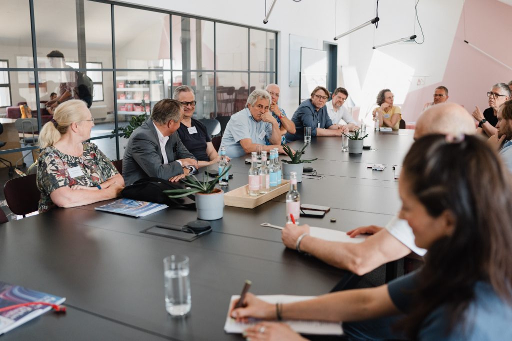 Mehrere Personen sitzen an einem langen Konferenztisch in einem modernen Besprechungsraum und führen eine Diskussion