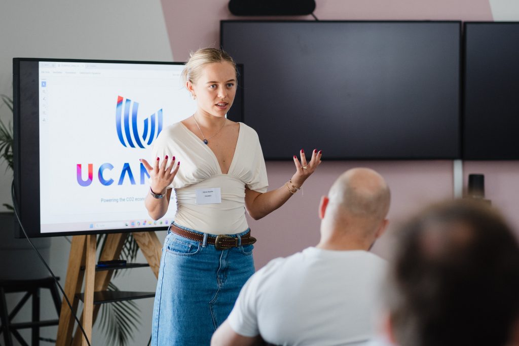 Junge Frau mit weißem Oberteil und Jeansrock spricht vor Publikum vor einem Bildschirm mit dem Logo 'UCANEO'