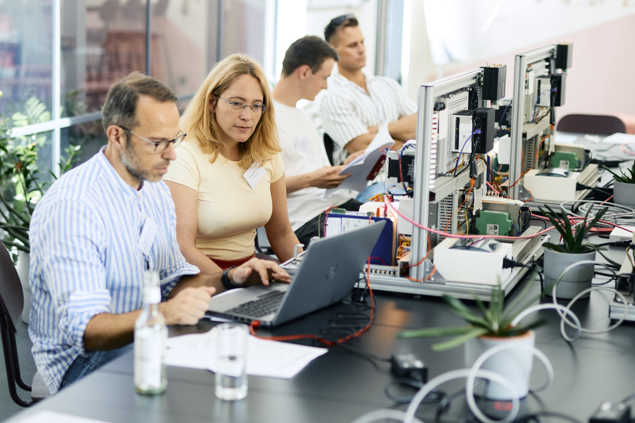Vier Personen sitzen an einem Tisch mit Laptops und technischen Geräten, zwei vorn konzentriert arbeitend, zwei hinten lesend und nachdenklich