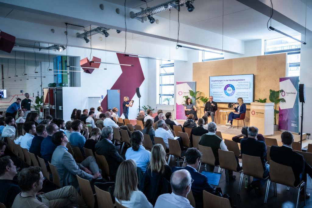 Publikum in einem modernen Veranstaltungsraum verfolgt eine Podiumsdiskussion mit drei Personen vor einer großen Präsentationsfolie