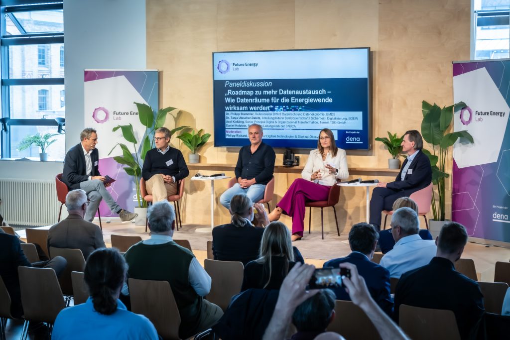 Fünf Personen sitzen auf einer Bühne vor Publikum und diskutieren in einem Panel, im Hintergrund ein Bildschirm mit Text und zwei Banner mit dem Schriftzug 'Future Energy Lab'